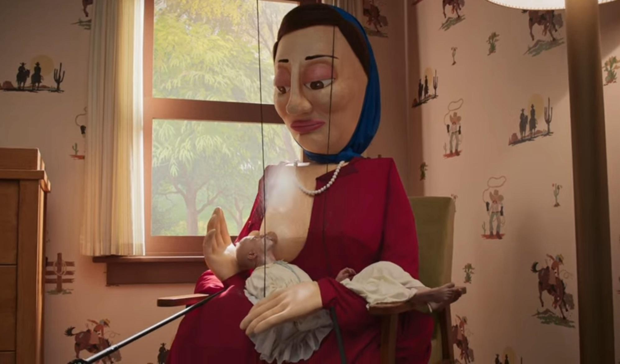 A large puppet figure of a woman in a red dress holding a baby, seated in a room with cowboy-themed wallpaper, a scene from The Rehearsal